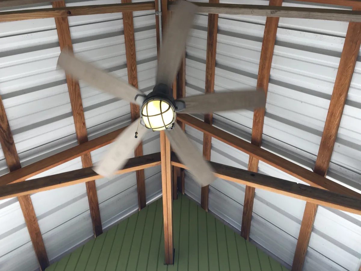 A ceiling fan with a caged light under the wood rafters of a metal-roofed pavilion